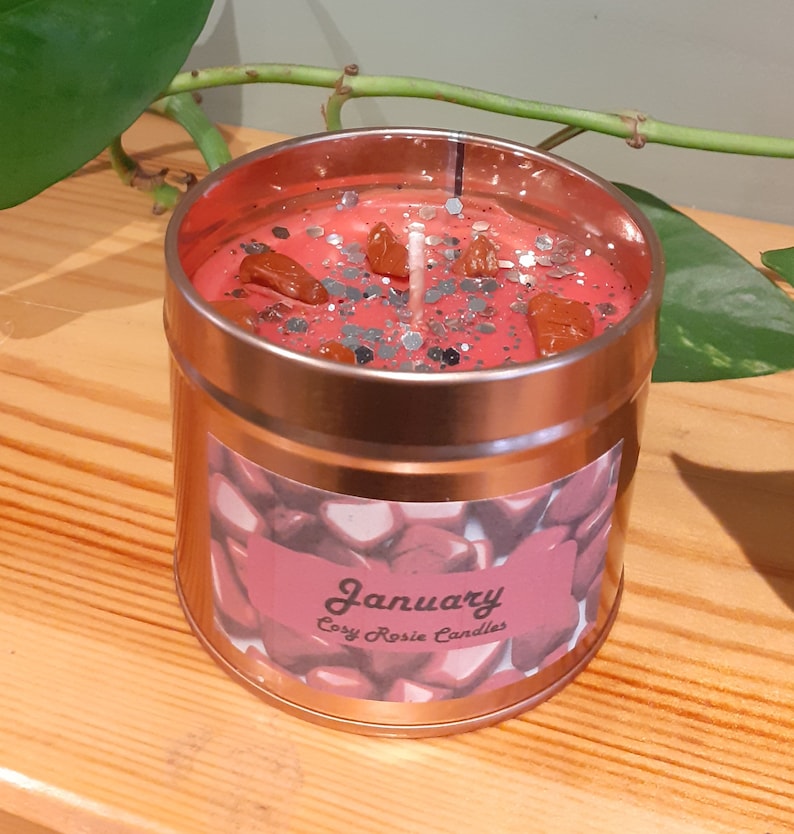 May include: A red candle in a copper tin with a silver glitter and brown crystal topping. The label reads "January Cozy Rosie Candles".