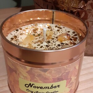 May include: A gold candle jar with a white lid and a label that says "November Cozy Rosie Candles". The candle is topped with a white wick and has a golden glitter and amber crystal topping.