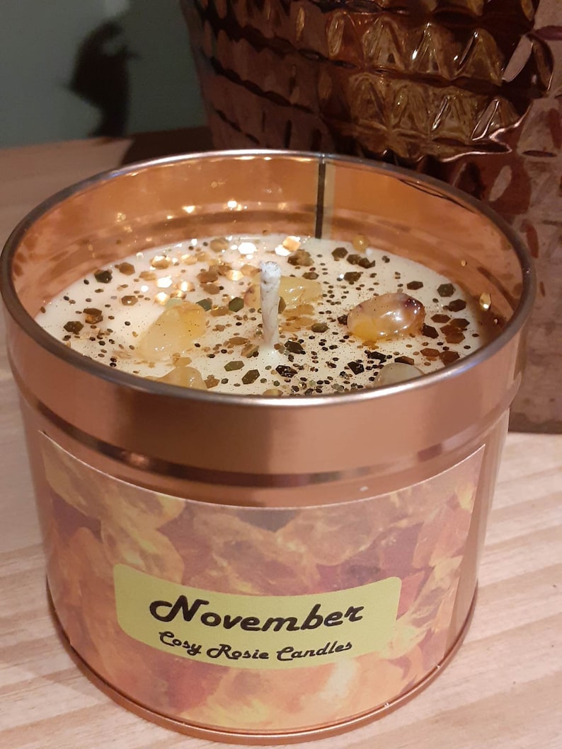 May include: A gold candle jar with a white lid and a label that says "November Cozy Rosie Candles". The candle is topped with a white wick and has a golden glitter and amber crystal topping.