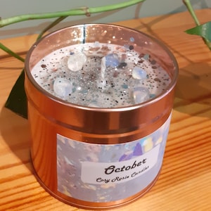 May include: A copper-colored candle jar with a white label that says "October Cozy Rosie Candles". The candle has a clear wax top with silver glitter and white stones.