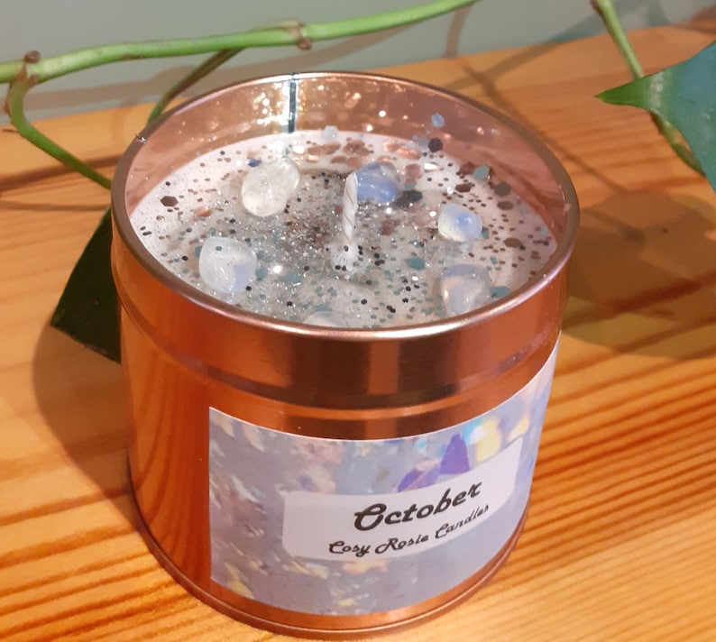 May include: A copper-colored candle jar with a white label that says "October Cozy Rosie Candles". The candle has a clear wax top with silver glitter and white stones.