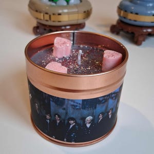 May include: A copper-coloured candle tin featuring a group photo. The candle has a dark wax base with glitter and three pink heart-shaped wax melts. A white wick is centered in the candle.
