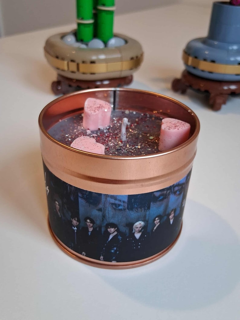 May include: A copper-coloured candle tin featuring a group photo. The candle has a dark wax base with glitter and three pink heart-shaped wax melts. A white wick is centered in the candle.