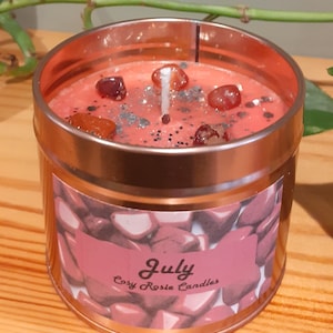 May include: A pink candle in a copper tin with a label that says "July Cosy Rosie Candles". The candle has red and orange crystals embedded in the wax.
