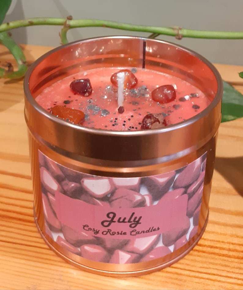 May include: A pink candle in a copper tin with a label that says "July Cosy Rosie Candles". The candle has red and orange crystals embedded in the wax.