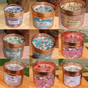 May include: Twelve candles in metal tins, each representing a different month of the year. The candles are decorated with various colors and glitter, and each tin has a label with the month's name and "Crystals & Herbs Candles".