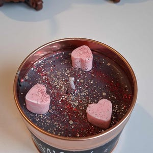 May include: A round candle in a copper-coloured tin. The candle wax is dark grey with red and silver glitter. Three pink heart-shaped wax melts are on top. The tin has black text that reads "Stray Kids".