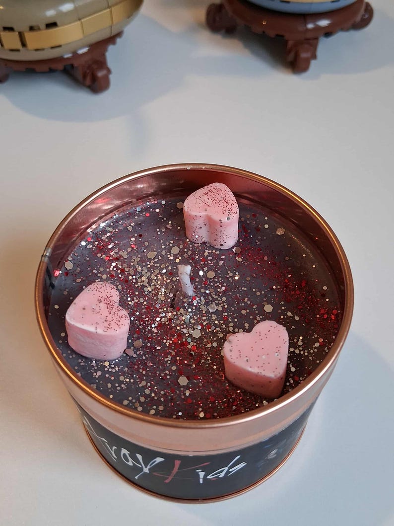 May include: A round candle in a copper-coloured tin. The candle wax is dark grey with red and silver glitter. Three pink heart-shaped wax melts are on top. The tin has black text that reads "Stray Kids".