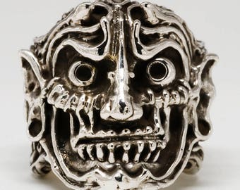 Può includere: Un anello d'argento con un design dettagliato e testurizzato di un volto demoniaco. L'anello presenta una fascia larga e un viso prominente, grottesco con denti aguzzi e corna.