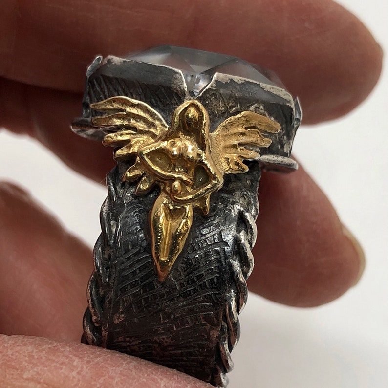 Puede incluir: Un anillo de plata con una gran piedra preciosa transparente y un dise&ntilde;o de &aacute;ngel de oro. La banda del anillo es texturizada y tiene un dise&ntilde;o trenzado.