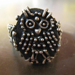 May include: A silver-toned owl ring with a detailed, textured design. The owl's face features large eyes and a small beak, with a body covered in small, raised dots. The ring's band is also adorned with similar textured details, creating a unique, handcrafted look.
