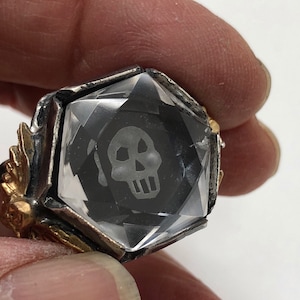 Puede incluir: Un anillo de plata con una piedra de cristal transparente engastada en una forma facetada. El cristal tiene un dise&ntilde;o de calavera negra en su interior. El anillo tiene una banda dorada.