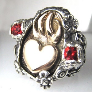 May include: A silver ring with a gold heart and flames, accented with red gemstones. The ring features a detailed floral design.