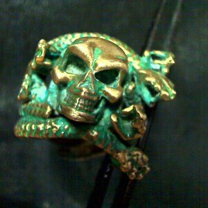 May include: A green and gold metal ring featuring a skull design with intricate details. The skull is surrounded by a snake-like design.