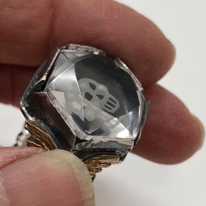 Puede incluir: Un anillo de plata con una piedra de cristal transparente engastada en un dise&ntilde;o facetado. La piedra de cristal tiene una calavera grabada en su interior. El anillo tiene detalles dorados.