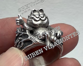Puede incluir: Un anillo de metal plateado que presenta una figura esculpida de un gato de dibujos animados con una amplia sonrisa y un gesto de pulgar hacia arriba, posado sobre una estructura similar a una rama. El texto @RXVrings 8/10/26 y RUBEN VIRAMONTES es visible.