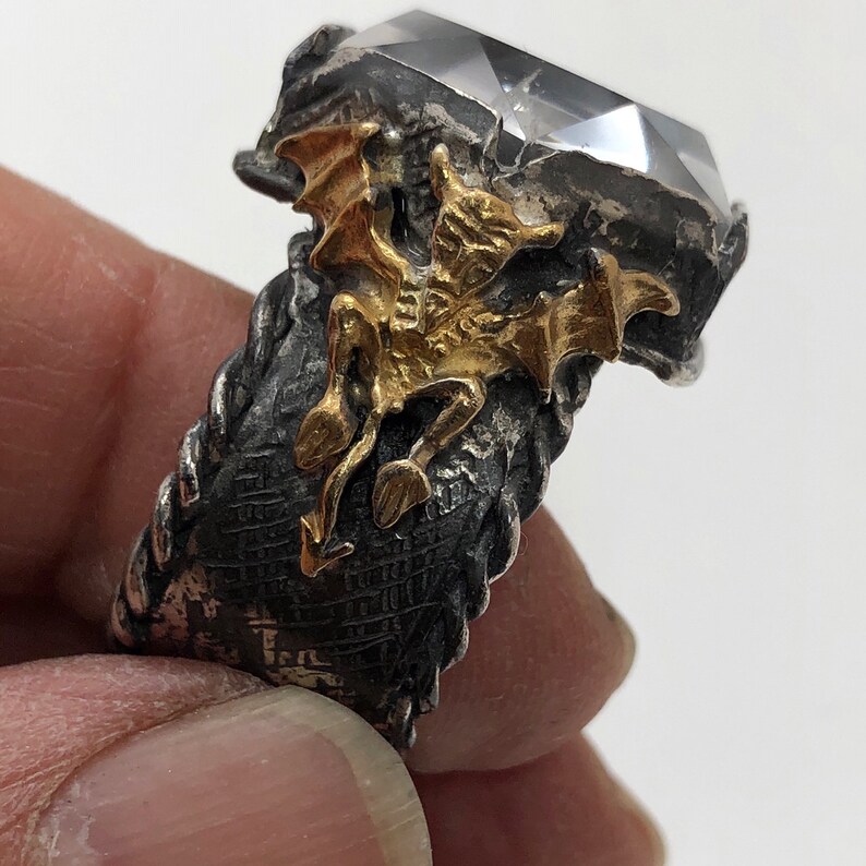 Puede incluir: Un anillo de plata con una piedra preciosa transparente y un dise&ntilde;o de drag&oacute;n dorado.