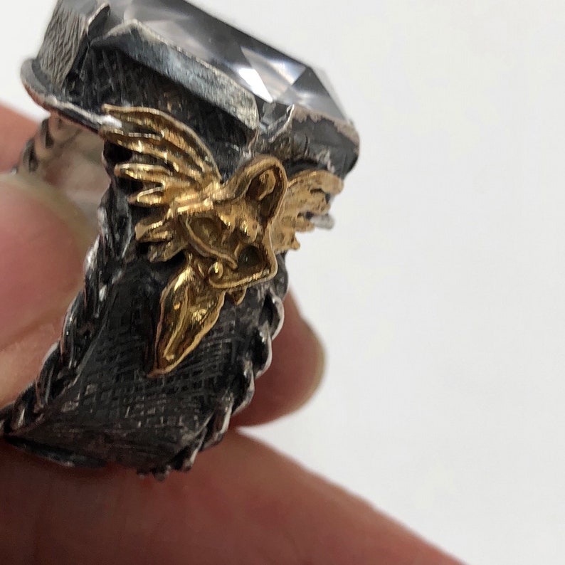 Puede incluir: Un anillo de plata con una gran piedra preciosa transparente y una figura alada dorada en el lateral.