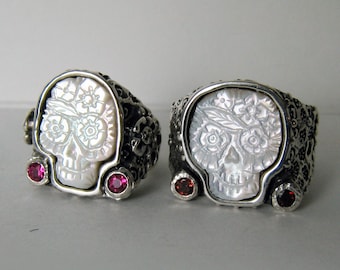 May include: Two silver rings with white skull designs and red gemstones. The skulls are carved with floral details and have a white mother-of-pearl finish.