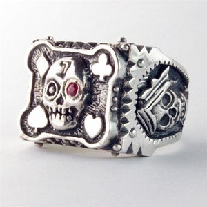 May include: A silver ring with a skull and playing card design. The skull has a red gemstone for an eye and is surrounded by playing card symbols. The ring has a textured, detailed design.