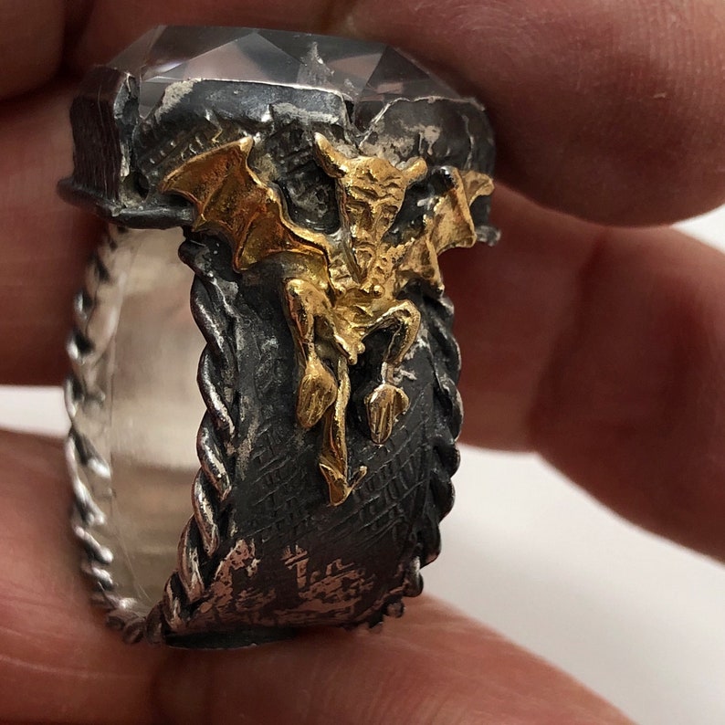 Puede incluir: Un anillo de plata con una banda negra y plateada y un dise&ntilde;o de g&aacute;rgola de color dorado. El anillo tiene una piedra transparente engastada en la parte superior.