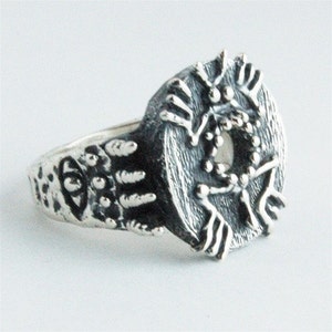 May include: A silver ring with an intricate design. The ring has a circular, textured centrepiece with a detailed pattern. The ring's band also features a textured design. The ring is set against a white background.