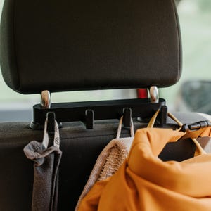 Op de afbeelding: Een zwarte kunststof hoofdsteun-organizer voor in de auto met vier geïntegreerde haken gemonteerd op de metalen steunstangen. De organizer houdt een grijs stoffen item en een oranje tas met een zwarte kunststof gespband vast.