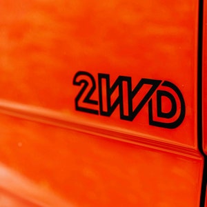 Op de afbeelding: Close-up van een feloranje oppervlak met de zwarte tekst "2WD" in een vetgedrukte, omlijnde lettertype. De afbeelding is goed belicht, waardoor het contrast tussen de kleuren en de strakke lijnen van de tekst wordt benadrukt.