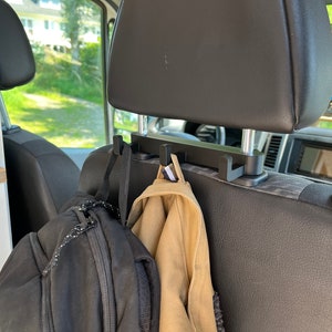 Op de afbeelding: Zwarte hoofdsteunhaken voor auto's met een rugzak en een beige jas die eraan hangen. De haken zijn bevestigd aan de hoofdsteun van een autostoel. De rugzak is zwart met een witte en zwarte touwritssluiting. De jas is beige.