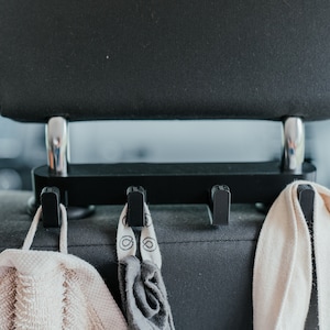 Könnte beinhalten: Schwarzer Haken für die Kopfstütze eines Autositzes mit drei Haken, an denen zwei weiße Stofftaschen und eine graue Stofftasche hängen.