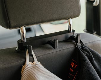 May include: A black plastic car headrest hook organizer featuring three individual hooks attached to the metal support posts of a vehicle seat. The device is mounted horizontally and holds a beige fabric item and a black fleece garment.