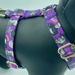 May include: Purple dog harness with a Halloween-themed pattern. The fabric features a repeating design of skulls, ghosts, and other spooky elements in black, white, and orange. The harness has silver metal hardware.