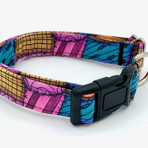 May include: A pet collar featuring a patchwork pattern of tan, pink, and blue fabric squares with black stitched details. The collar includes a black plastic side-release buckle and a silver-toned metal D-ring for attaching a leash or identification tag.