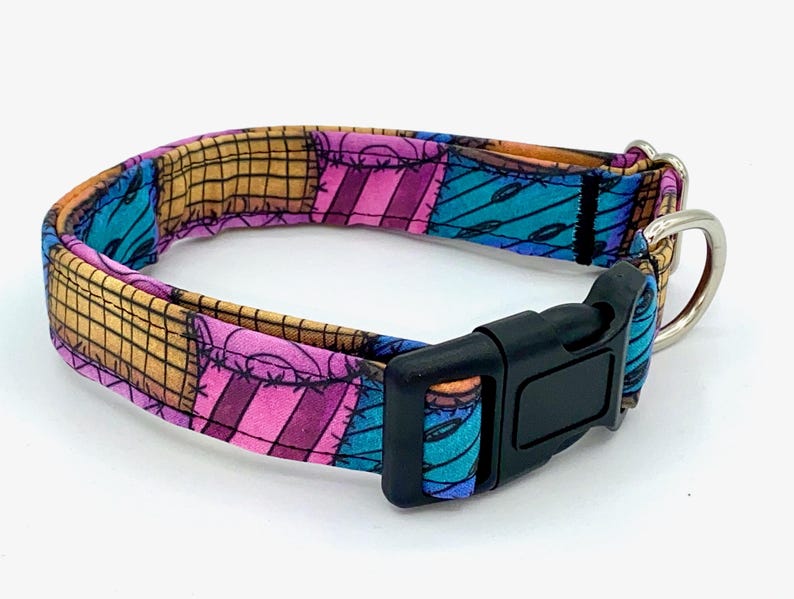 May include: A pet collar featuring a patchwork pattern of tan, pink, and blue fabric squares with black stitched details. The collar includes a black plastic side-release buckle and a silver-toned metal D-ring for attaching a leash or identification tag.
