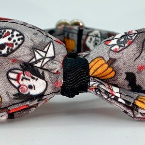 May include: Gray Halloween-themed bow tie with a black center. The fabric features a pattern of pumpkins, bats, and horror movie characters. The bow tie has a silver clasp.