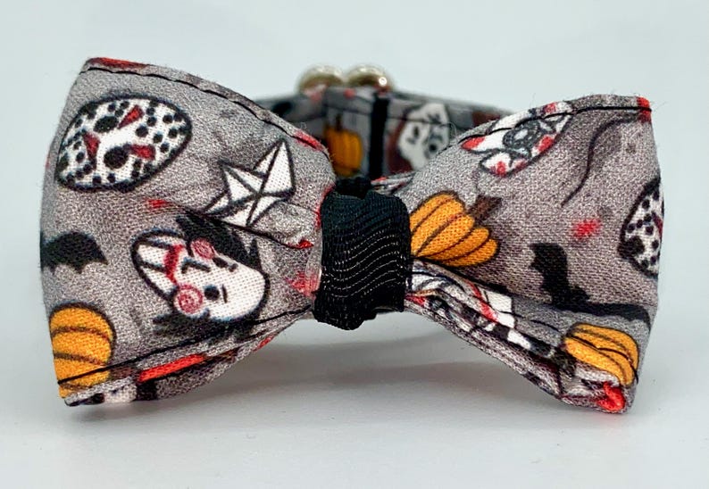 May include: Gray Halloween-themed bow tie with a black center. The fabric features a pattern of pumpkins, bats, and horror movie characters. The bow tie has a silver clasp.