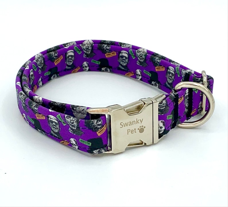 May include: A purple dog collar featuring a repeating pattern of classic monster faces in black and white. The collar has a silver metal buckle and a D-ring for leash attachment. The brand name "Swanky Pet" is printed on the buckle.