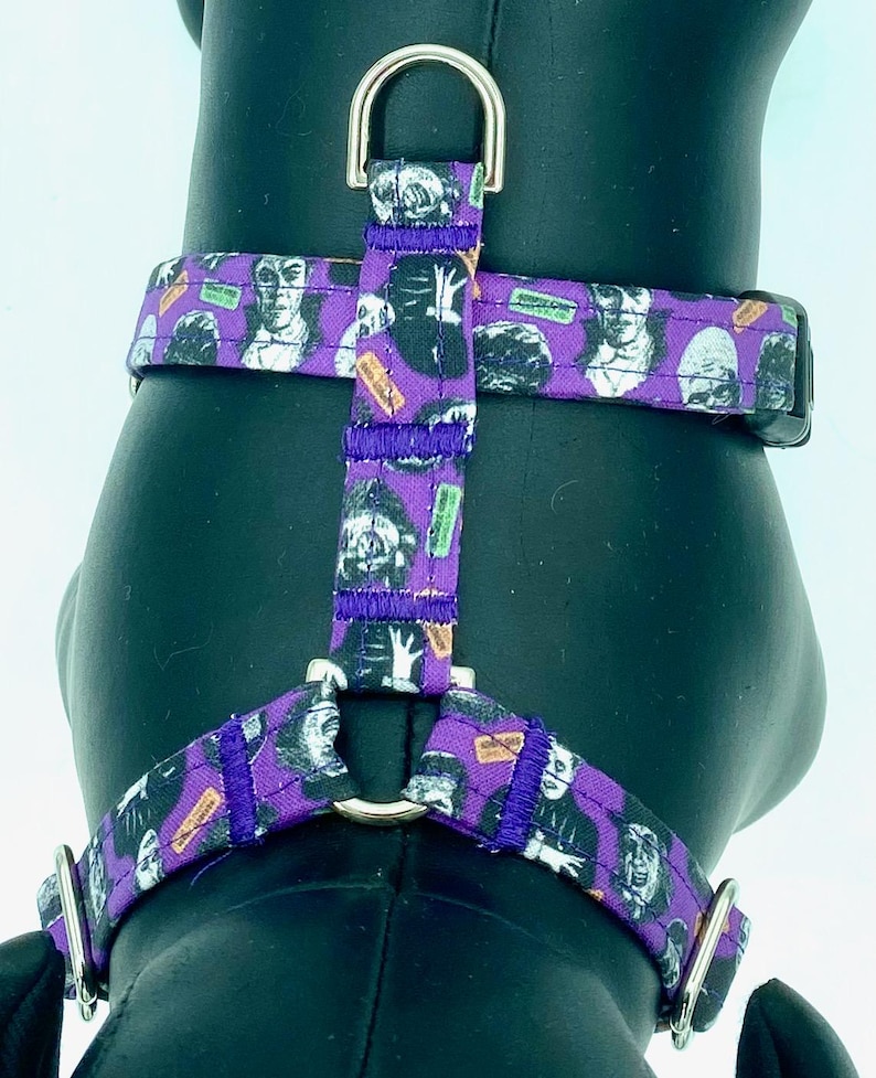 May include: Purple dog harness featuring a repeating pattern of classic horror movie monsters in black and white. The harness has a metal D-ring for leash attachment and adjustable straps. The harness is designed for a comfortable and secure fit.