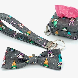 May include: A gray fabric set featuring a wristlet, bow tie, and a small bag with a zipper and a carabiner clip. The items are decorated with a repeating pattern of colorful gnomes and bunnies on a dotted background.
