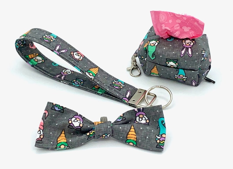 May include: A gray fabric set featuring a wristlet, bow tie, and a small bag with a zipper and a carabiner clip. The items are decorated with a repeating pattern of colorful gnomes and bunnies on a dotted background.