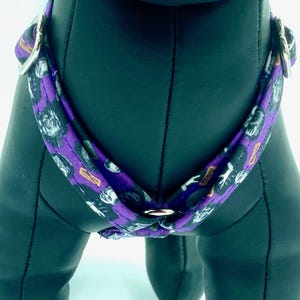 May include: A purple dog harness with a repeating pattern of black and white skulls and bones. The harness has silver metal hardware and is designed to fit around the dog's chest and shoulders.