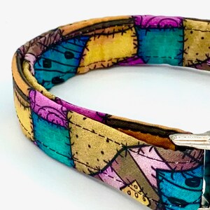 May include: A fabric collar featuring a patchwork design with various colored squares in shades of teal, purple, yellow, and brown. The pattern includes black stitched lines and small dot details, finished with a metallic silver buckle closure.