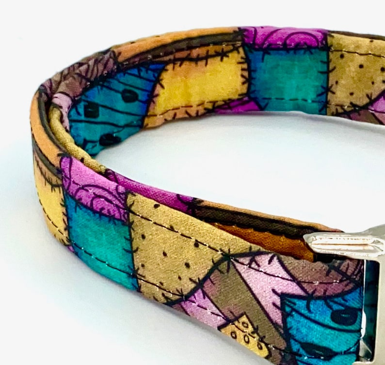 May include: A fabric collar featuring a patchwork design with various colored squares in shades of teal, purple, yellow, and brown. The pattern includes black stitched lines and small dot details, finished with a metallic silver buckle closure.