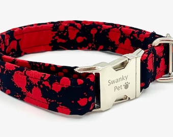 May include: A dog collar with a red and black splatter pattern. The collar has a silver-colored metal buckle and a D-ring. The text "Swanky Pet" is printed on the buckle.