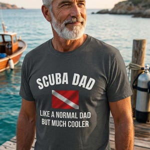 以下が含まれることがあります： 「SCUBA DAD」の文字と赤と白のダイビングフラッグのグラフィックが入った濃いグレーのTシャツ。シャツには「LIKE A NORMAL DAD BUT MUCH COOLER」とも書かれています。画像には、ボートとスキューバダイビングのギアの近くの木のドックにいる男性が写っています。