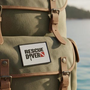 Puede incluir: Una mochila verde oliva con correas y detalles de cuero marrón. La mochila presenta un parche rectangular con las palabras "RESCUE DIVER" en negro y rojo. La bolsa tiene múltiples compartimentos y hebillas metálicas.