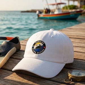 Puede incluir: Una gorra de béisbol blanca con un parche circular que muestra una tortuga marina y el texto Bonaire Scuba Diving, acompañada de una aleta de natación negra y beige, un esnórquel negro y una brújula de latón sobre un muelle de madera.