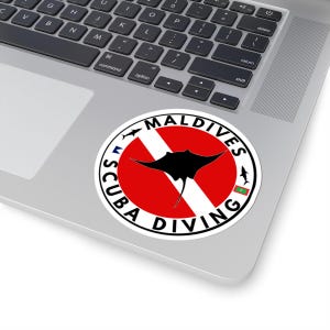 Puede incluir: Una pegatina redonda con las palabras "MALDIVES SCUBA DIVING". El diseño incluye una manta raya negra sobre un fondo rojo y blanco, con pequeños peces y la bandera de Maldivas. La pegatina está sobre un portátil.
