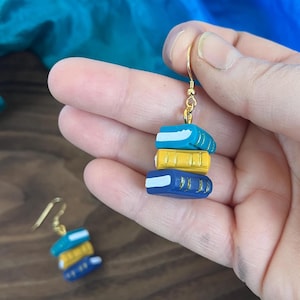 May include: Gold-tone earrings featuring a stack of three miniature books. The books are painted in blue, yellow, and dark blue with white accents. The earrings are held in a hand against a teal and blue background.