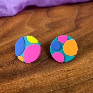 May include: Two colorful, round stud earrings. Each earring features a teal base with overlapping shapes in pink, blue, yellow, and orange. The earrings are displayed on a wooden surface.